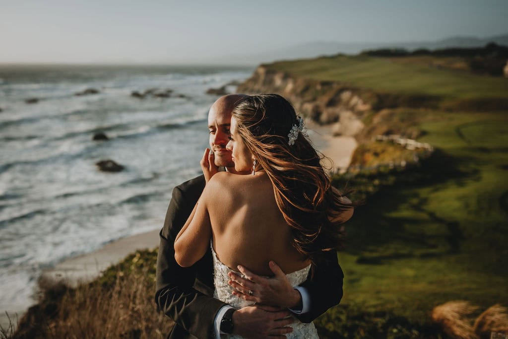 Bride and groom embracing on a scenic coastal cliff at sunset.