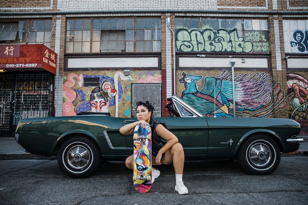 Young woman with skateboard and vintage car in urban graffiti setting.